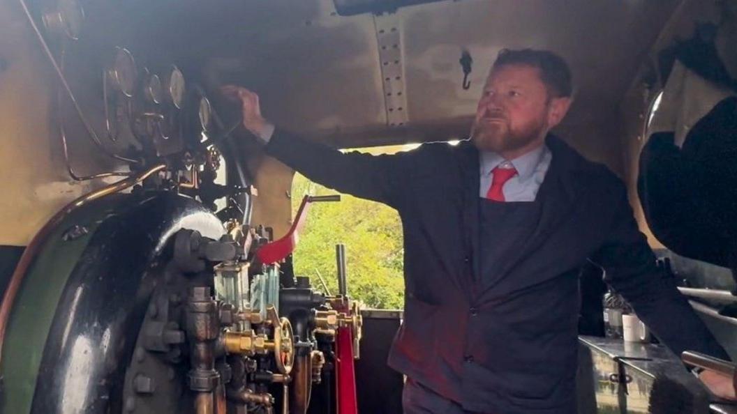 A steam locomotive driver sounding the whistle from inside the cab. He has a beard and is wearing a blue uniform with a red tie.