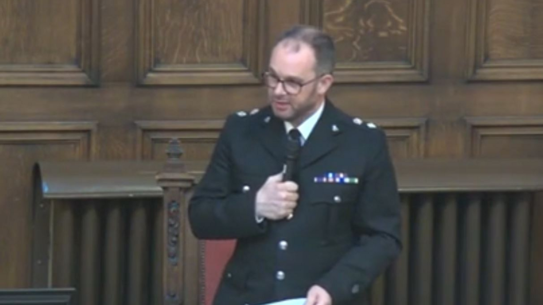 A police officer in formal uniform holding a microphone and addressing a room.