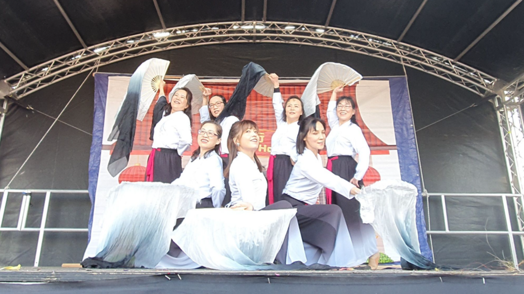 Seven female Chinese dancers dressed in white shirts and long black skirts. They are waving white fans which are covered in scarves and they are all performing on a stage.