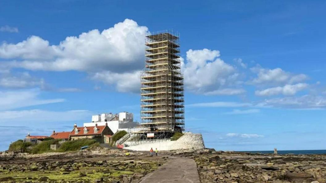 A view of St Mary's Island. A causeway leads to the scaffolded lighthouse, which is surrounded by several houses. Two people are walking across the causeway. The weather is sunny. There are rocks on both sides of the causeway. 