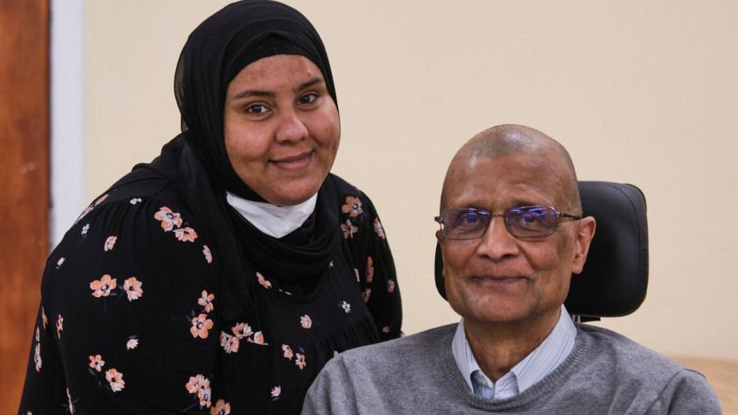 A younger woman (on the left) is standing and smiling next to an older man (on the right), who has a head rest behind him. The woman is smiling and wearing a black top with pink flowers and a black hijab. The man is smiling and wearing glasses, a light coloured shirt and a grey jumper. Behind them is a plain wall.  