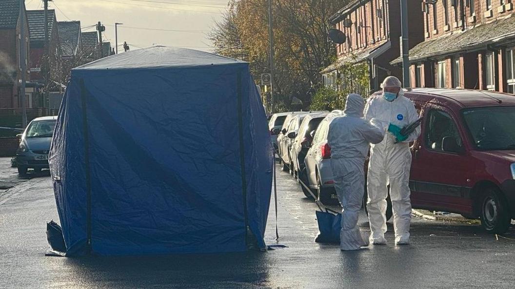 Two scene of crime officers, wearing white suits and blue gloves, are photographed in conversation as they stand next to a blue forensics tent that has been erected in the middle of the road.