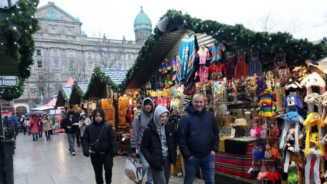 People walking in the ground of Belfast City Hall. Wooden huts line the pavement covered in garlands and lights. 