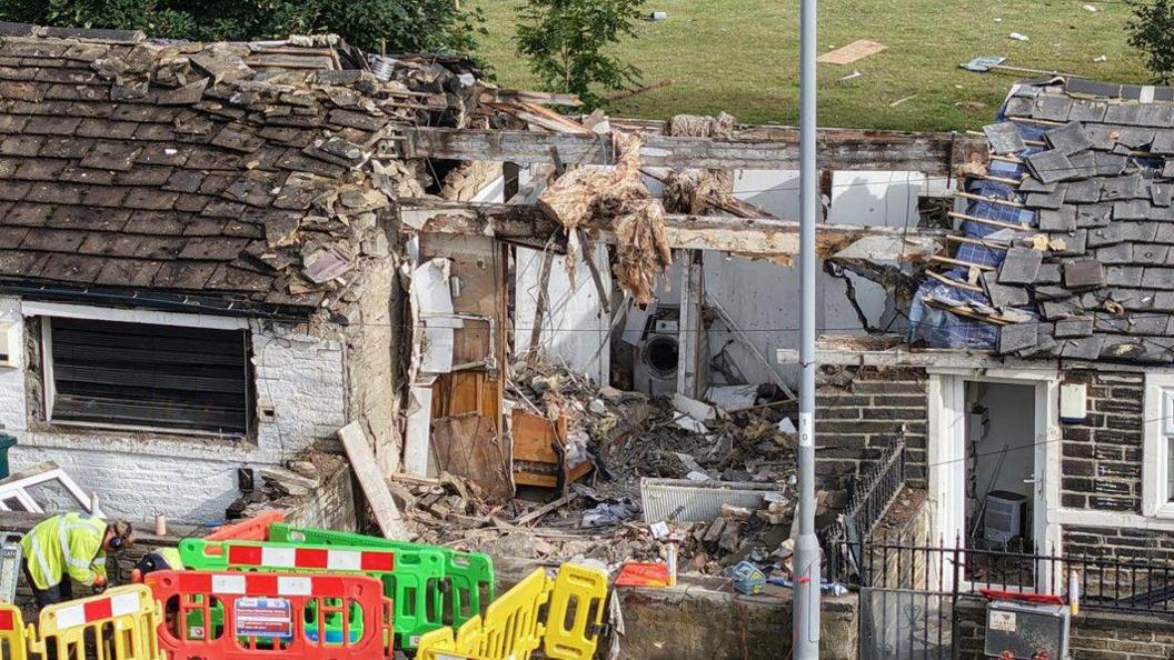 An close up shot of a terraced house with extensive explosion damage including a decimated roof and debris all over the road and back garden