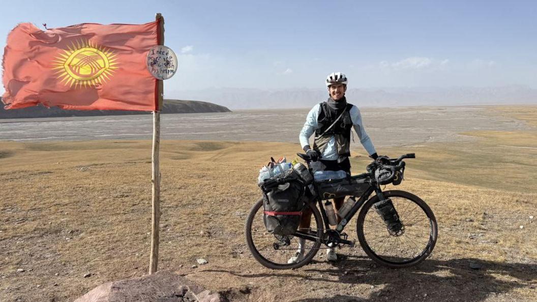 Finlay Tusting, wearing a cycle helmet, scarf, long-sleeved top and gloves stands behind his bike next to a flag flying from a wooden pole. The terrain is rough, with mountains in the background. There is no-one else around.