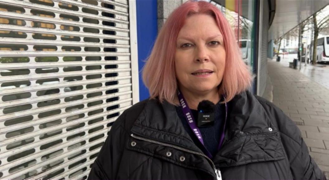 A woman with pink hair stands in front of a shopfront on a city street. A coach is visible in the background. 