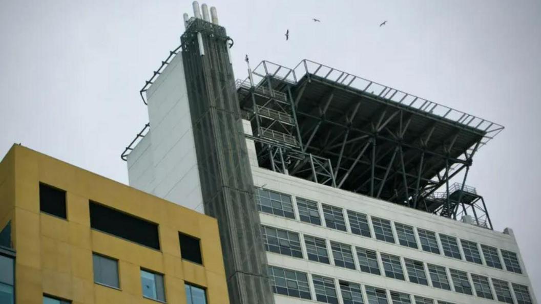 A metal platform on top of a white building, there is a yellow building to the left of it and a grey sky beyond