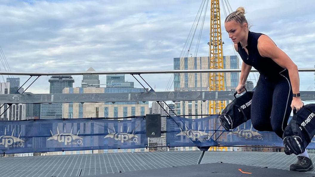 A woman lifts two sandbags and runs with them at the top of an arena in London