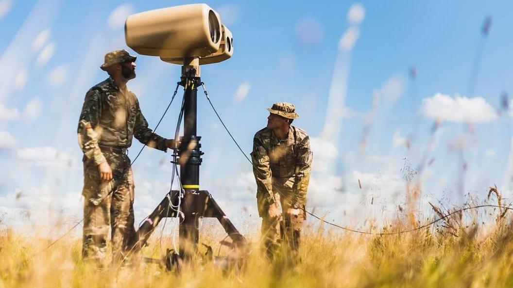 Two men in military khaki use a bit of kit that is on a stand