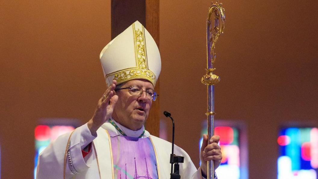 John Sherrington wears an ornately decorated white and gold mitre and a white and lilac robe. He holds a long, thin staff in his left hand known as a crosier.