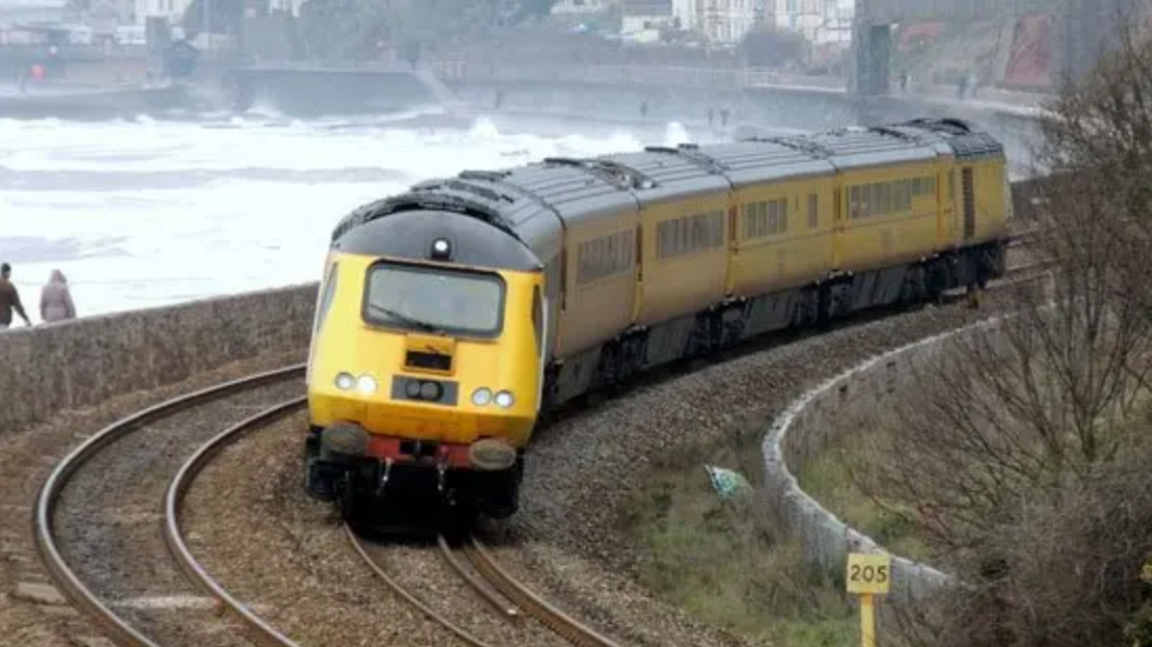 A yellow train coming round a bend on a track. To the left of it, is the sea and coast with two people walking. A cliff edge can be seen in the distance above the sea with houses at the top.