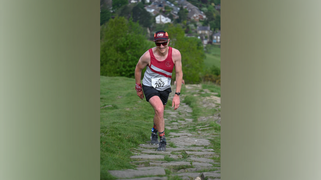 A man wearing a running vest with a number on it, and a cap and sunglasses, walks up a large stoned hill with grass either side 