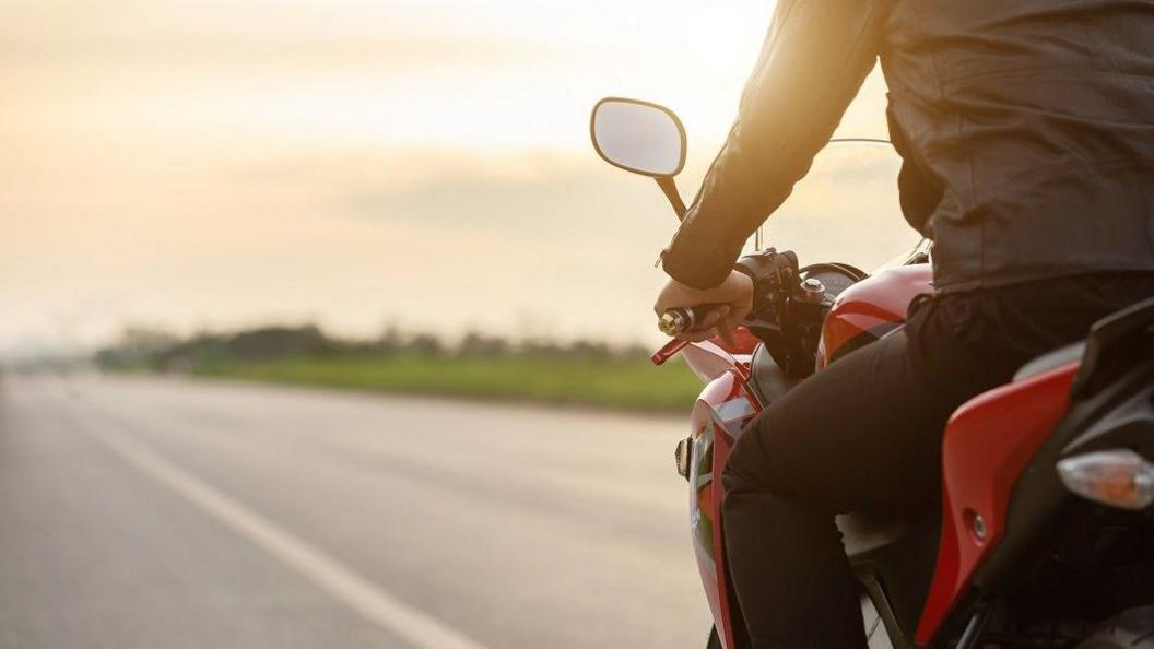 A motorbike rider with a dark jacket and dark trousers, sits on a red motorbike on the edge of the photo. Ahead of them we can see a road with a grass verge to one side with a hint of sunshine above.