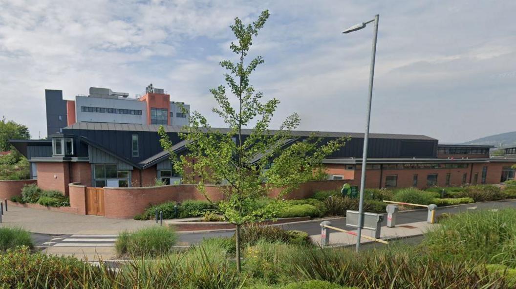 A one storey red brick building with a black roof. Belfast City Hospital is visible behind the building. In front of it is bushes and a tree. 