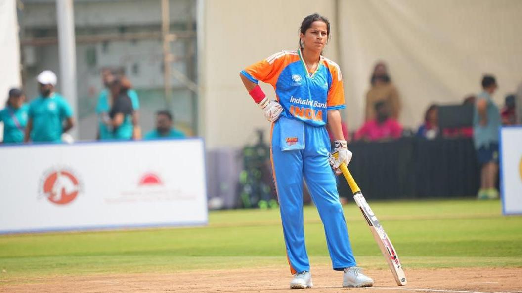Batter Anekha Devi in her blue and orange jersey, pictured on the field, holding her bat. 