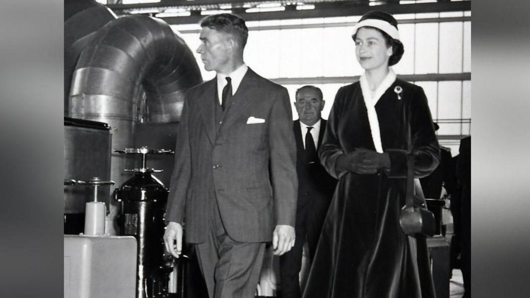 A black and white image of a young Queen Elizabeth II walking inside Calder Hall next to a man in a suit with a white handkerchief peeping out of his top pocket. The Queen is wearing a dark velvet dress and matching hat. A larger, bald man is behind her and her companion, seen through the gap between them.