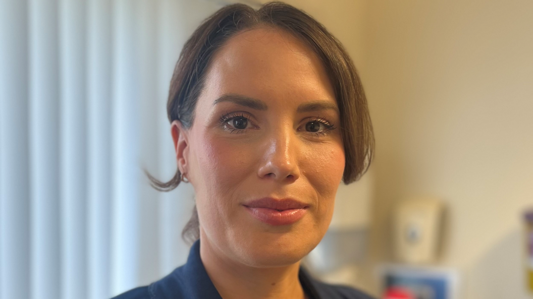 Dr Jilly O'Hagan has brown hair parted in the middle and tied back in a pony-tail and has brown eyes. She is wearing a navy suit jacket