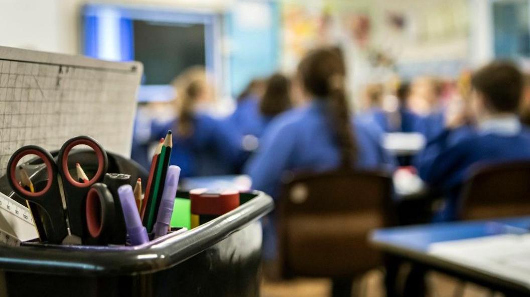 Children in a blue school uniform are sitting at their desks in a classroom. Their images are blurred but in the foreground, a box of stationary is in focus.