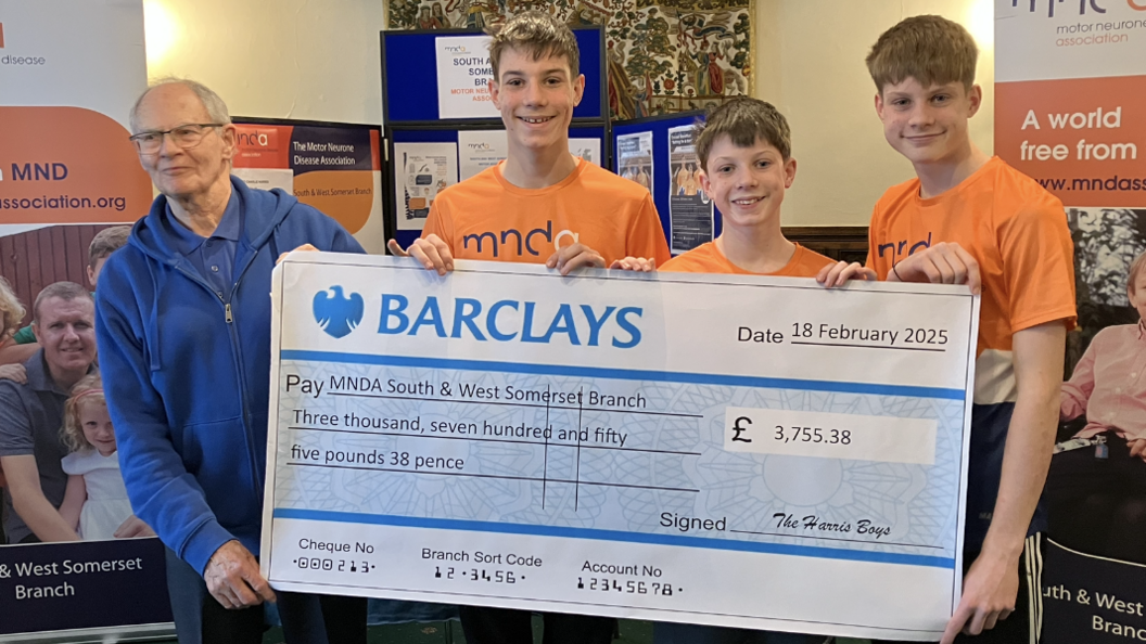 Three boys wearing orange tshirts present a cheque to a man in a blue jacket.