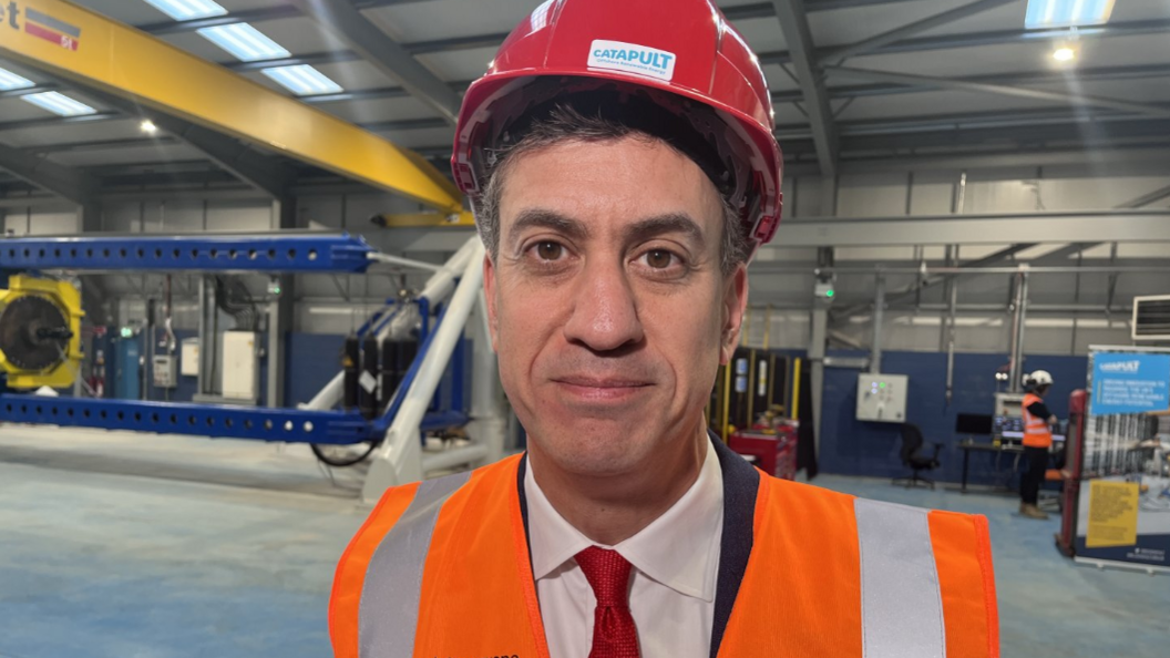 Energy secretary Ed Miliband wearing a red hard hat and orange hi-vis vest, smiling at the camera. 