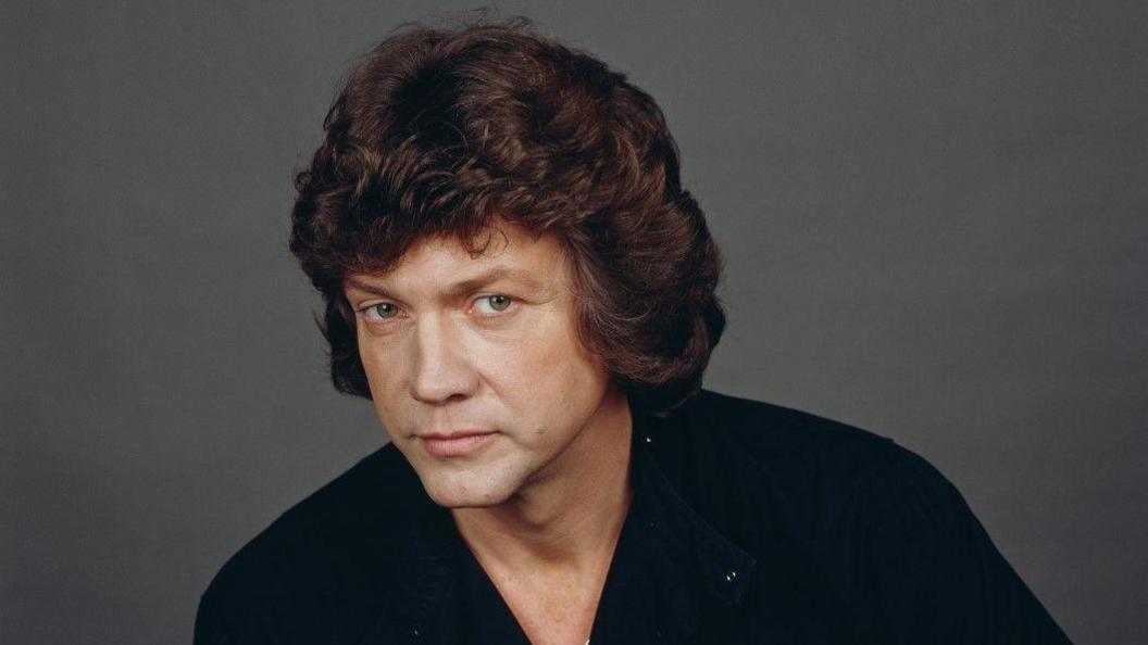 A man with curly brown hair is looking at the camera. He is wearing a dark top.
