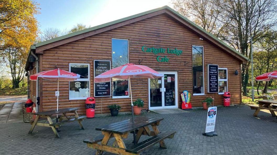 Cartgate Lodge Cafe. It is a large wooden clad shed with tall windows. There are three picnic benches and parasols outside on the paved area, and trees dotted around.