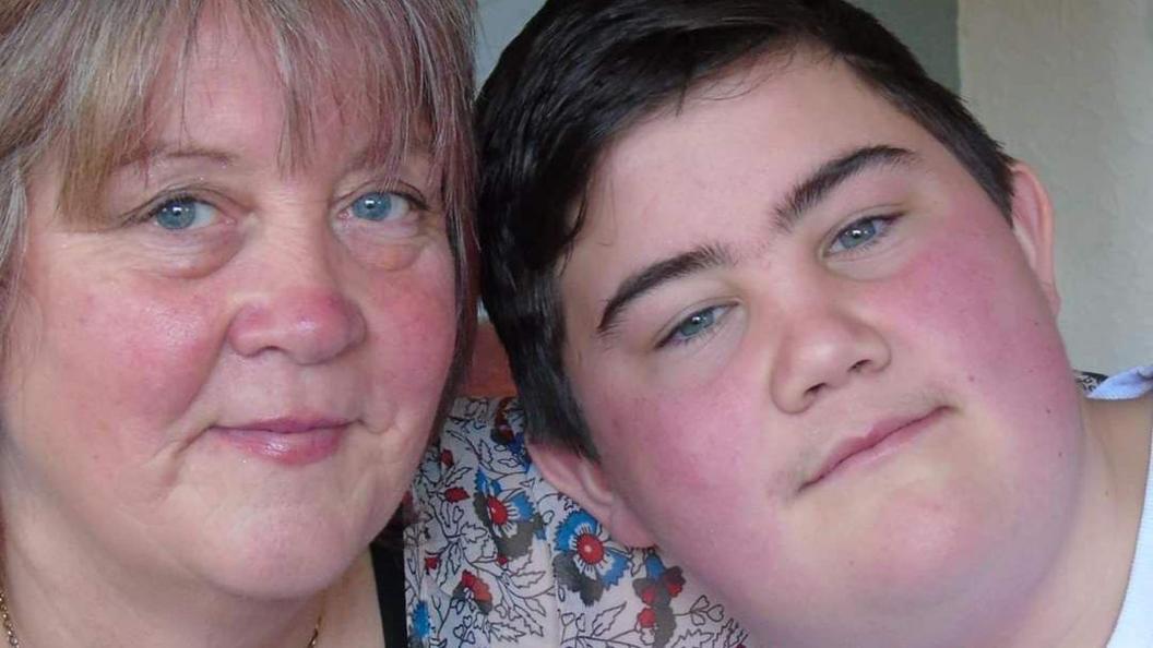 A woman and a boy pose for a photo, the woman is wearing a floral patterned top.