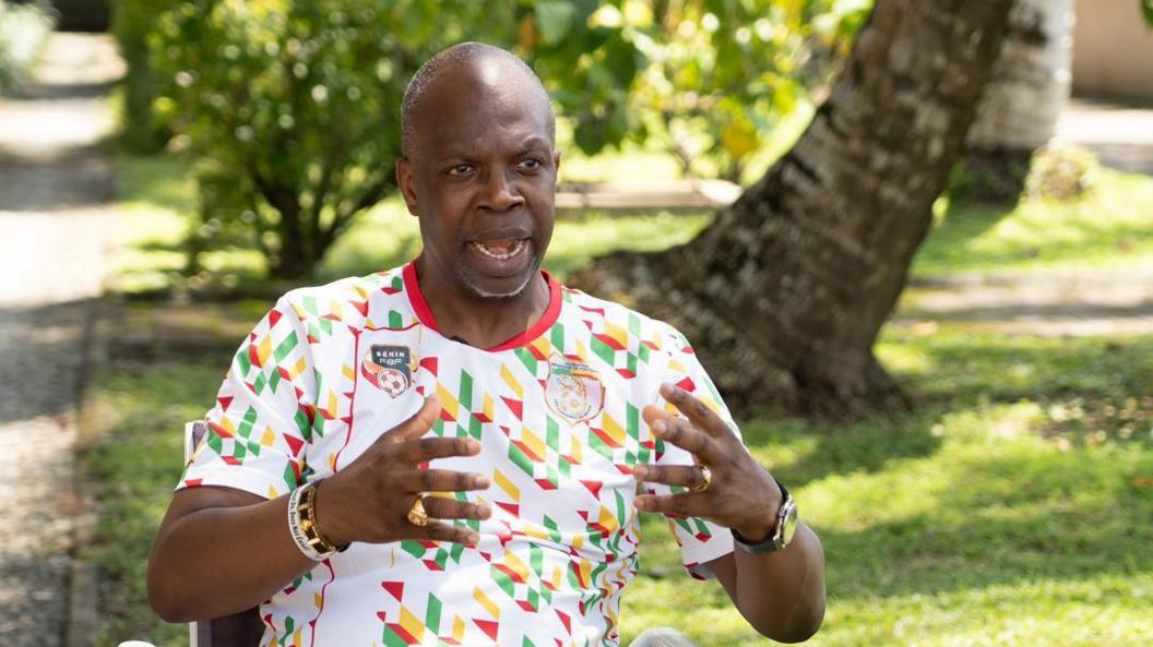 Tunde Adelakun wearing a Benin training kit, sitting with his mouth and hands opened