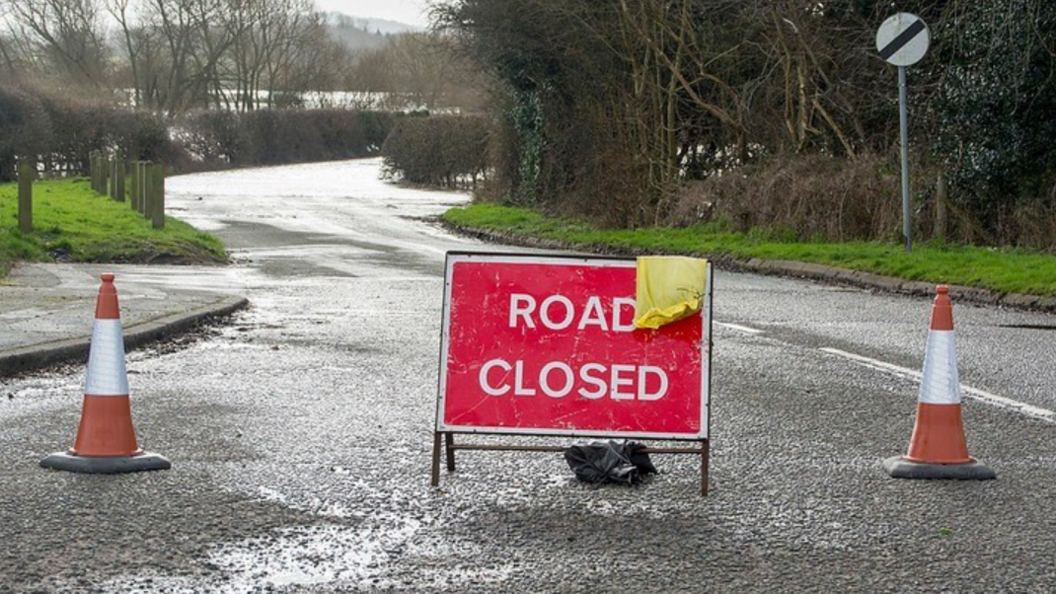 A red road closed sign with a yellow cloth hanging from the top on a wet road, with a traffic cone on either side and flooding further down the road