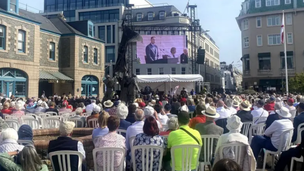 People are sat down at a previous event. They are sitting down on white chairs and looking at a large screen. There is buildings pictured around the side with a white tent at the front.
