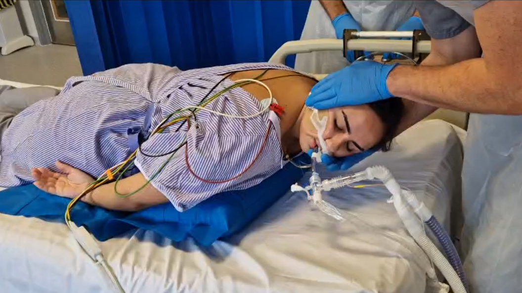 A female patient is laying on her chest, seemingly asleep, on a hospital bed. She is in a medical room with dark blue curtains separating patient beds. The patient has a medical tube attached to her nose. She is laying over a blue pillow-like device that is slowly being inflated, while a medical professional supports the patient's head.