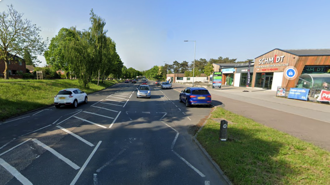 Cars travelling along Hall Road. A grass bank and trees line one side of the street, a retail park with wood-clad buildings lines the other