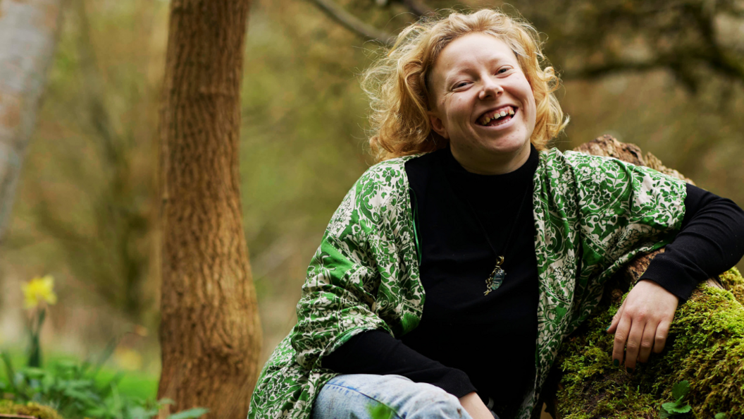 Athena Aperta sat on a log in the woods. There are trees around her. She is smiling and wearing a green shirt over a black top.