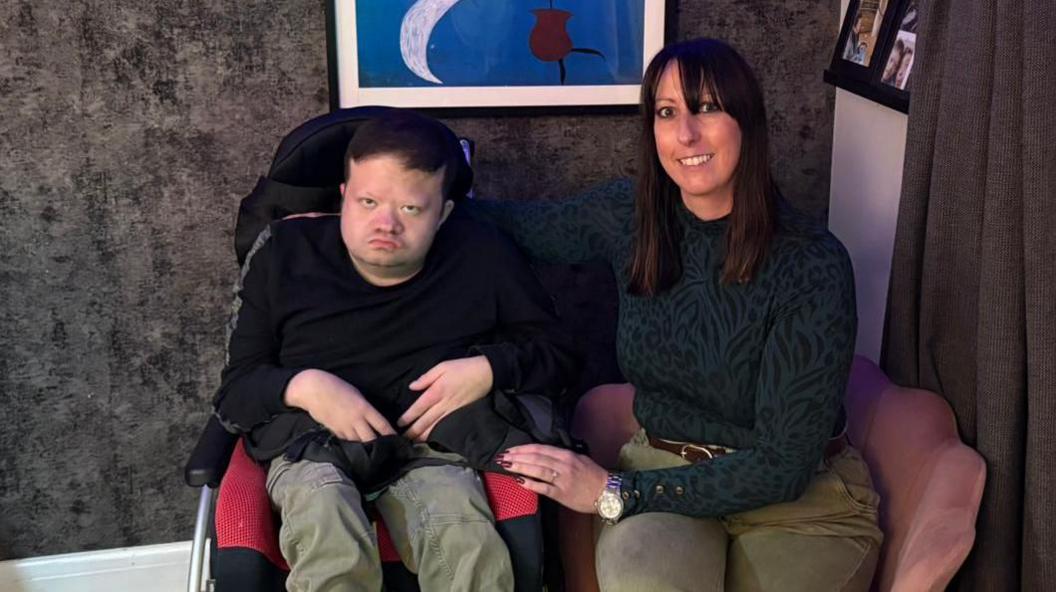 A young man sits in a wheelchair looking at the camera. A woman is sitting next to him on an armchair, with her arm around him. She is smiling at the camera.