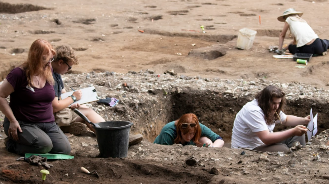 Students and staff in the trenches digging with tools, some sitting on the ground with notepads making notes of their findings