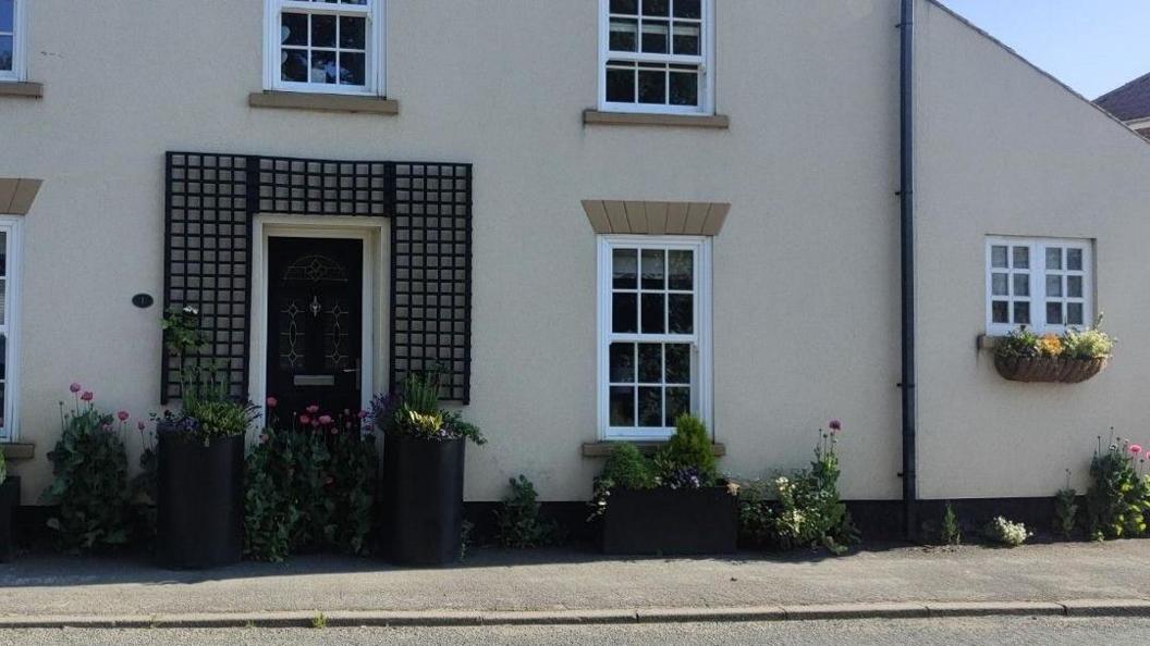 The front of a beige house with a black front door at the side of a road. You can see two tall planters with flowers on each side of the front door, a shorter but longer one underneath a window and more greenery and flowers surrounding them. There is a footpath in front of them.