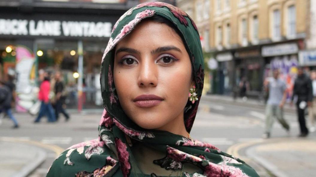 Nabeela Zaman wearing a green and pink floral hijab, in the background is a high street area, blurred out slightly.