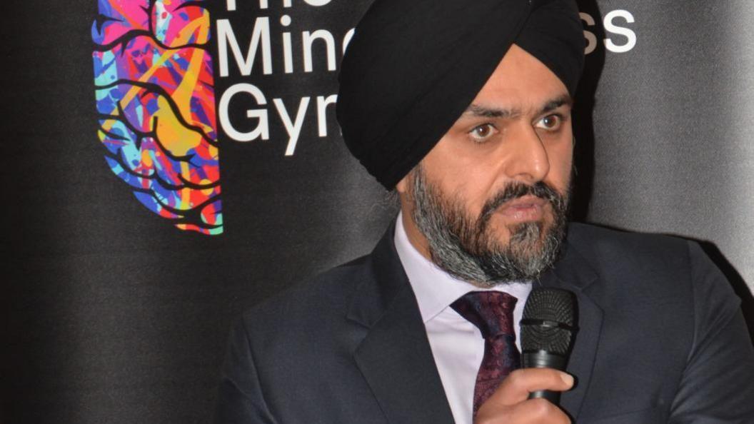 Bhupinder Sandhu speaking at a public event, holding a microphone. He wears a dark suit, white shirt, dark red tie and a navy blue turban.