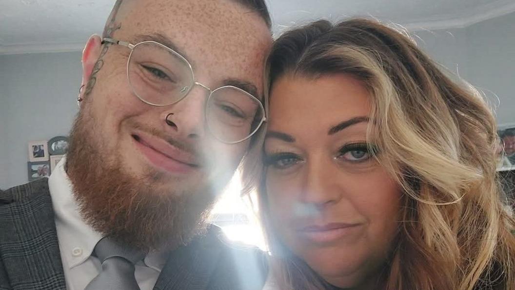Connor, who has a ginger beard and silver-rimmed glasses, is wearing a white shirt, grey tie and grey checked waistcoat. He is smiling at the camera and posing with his mother Toni, who has shoulder-length blonde hair.