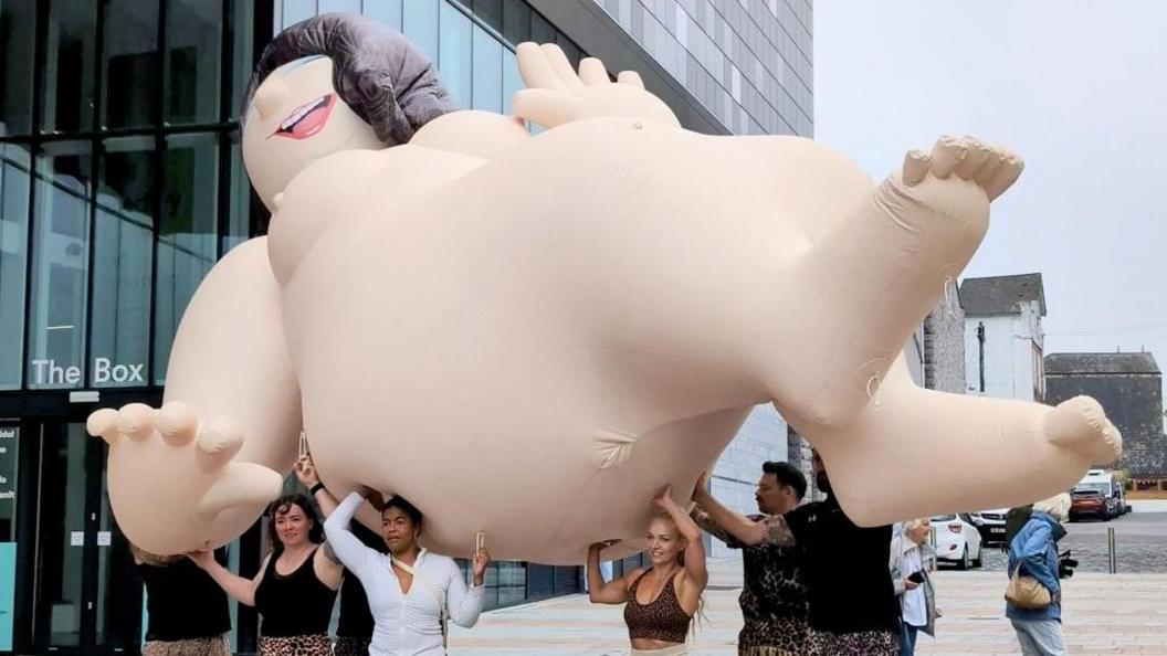 A naked inflatable character being carried by five strong people outside The Box in Plymouth.