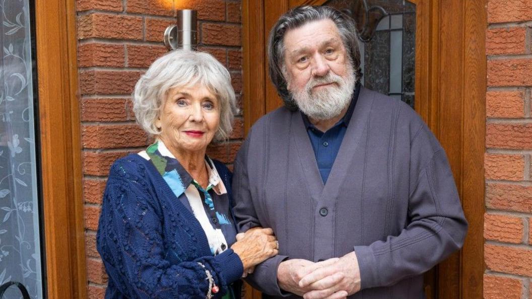 Sue Johnston and Ricky Tomlinson are pictured in character standing in the doorway of a red-bricked home.