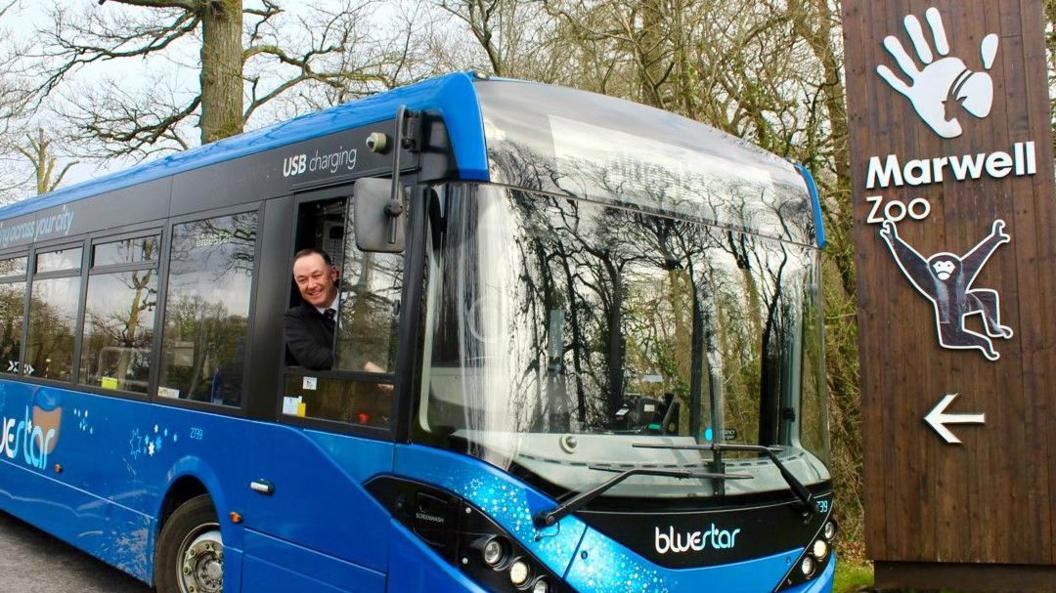 Brown sign for zoo on right with monkey logo and blue bus to the left with driver at the window.