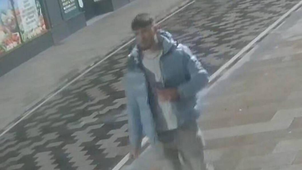 A man wearing a light blue puffer jacket, light jeans, a white top and a necklace, on an empty street. The image is blurry and taken by a CCTV camera.