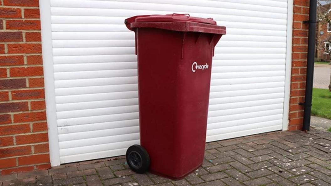 A burgundy recycling bin on pavement in front of a white garage.