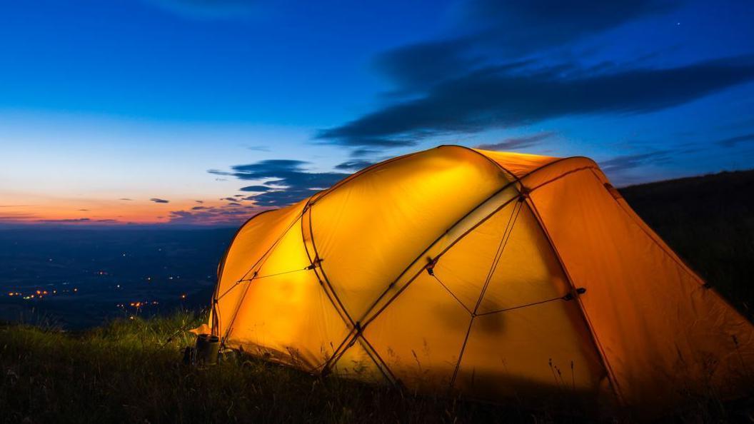 A yellow tent lit from inside. It is on a patch of grass. Lights can be seen in the distance. The sky is blue and orange with some clouds.