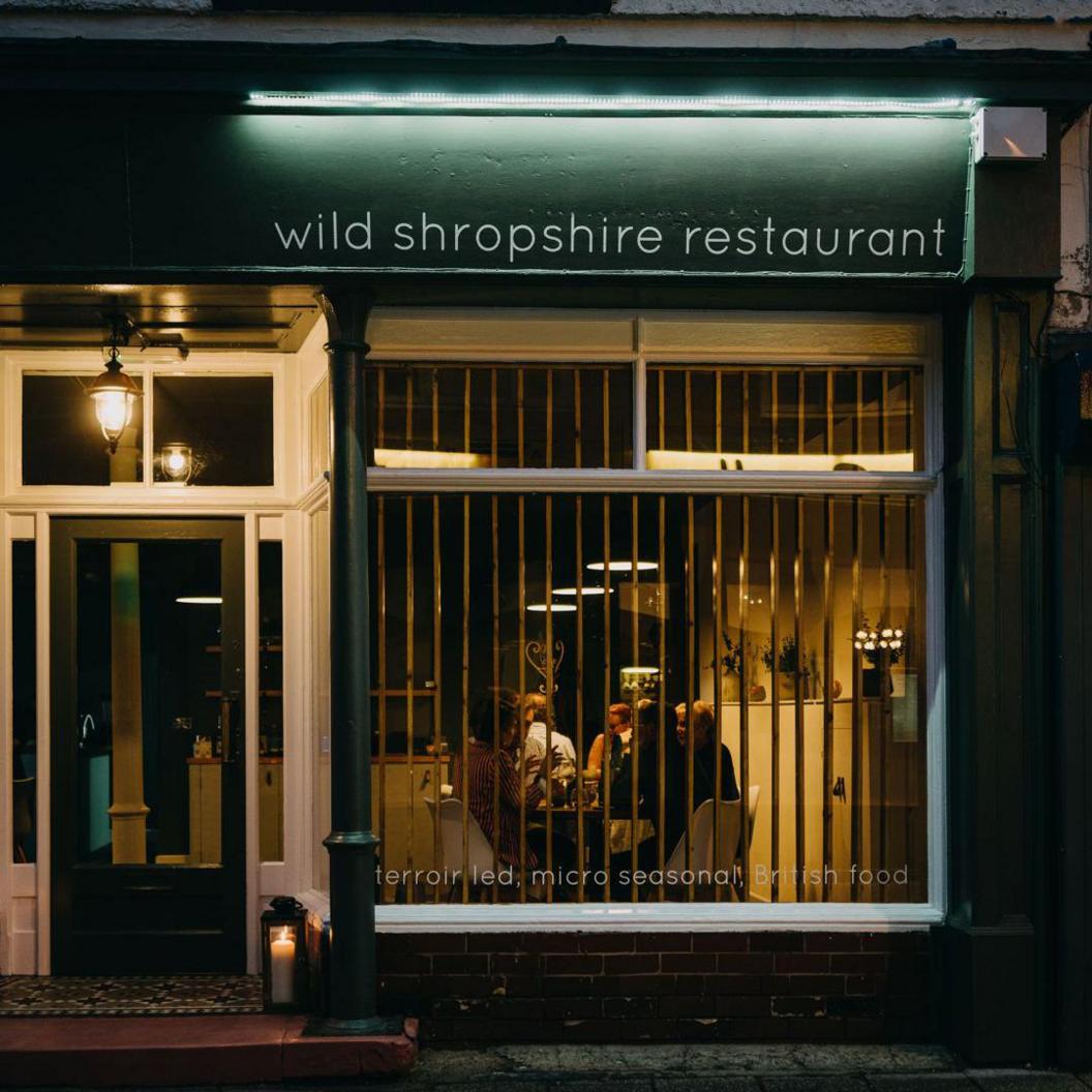 The front of a restaurant with a front door on the left and people sitting inside at tables visible through the window. A green sign is over the window with "Wild Shropshire restaurant" in white letters.