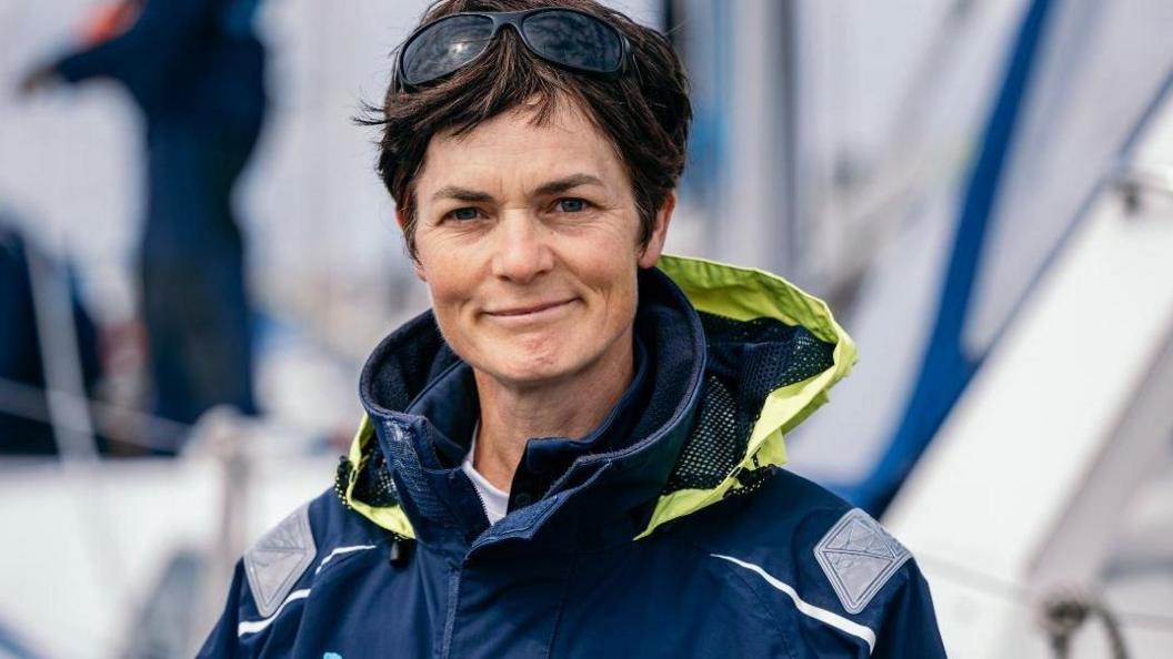 Dame Ellen MacArthur wearing a blue sailing jacket looking straight at the camera.  She has short brown hair and sunglasses on top of her head.