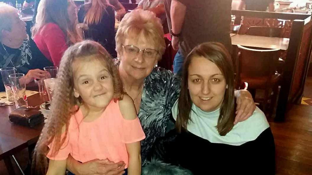 Older woman with glasses with her hands around a woman with dark hair and a girl with blonde hair