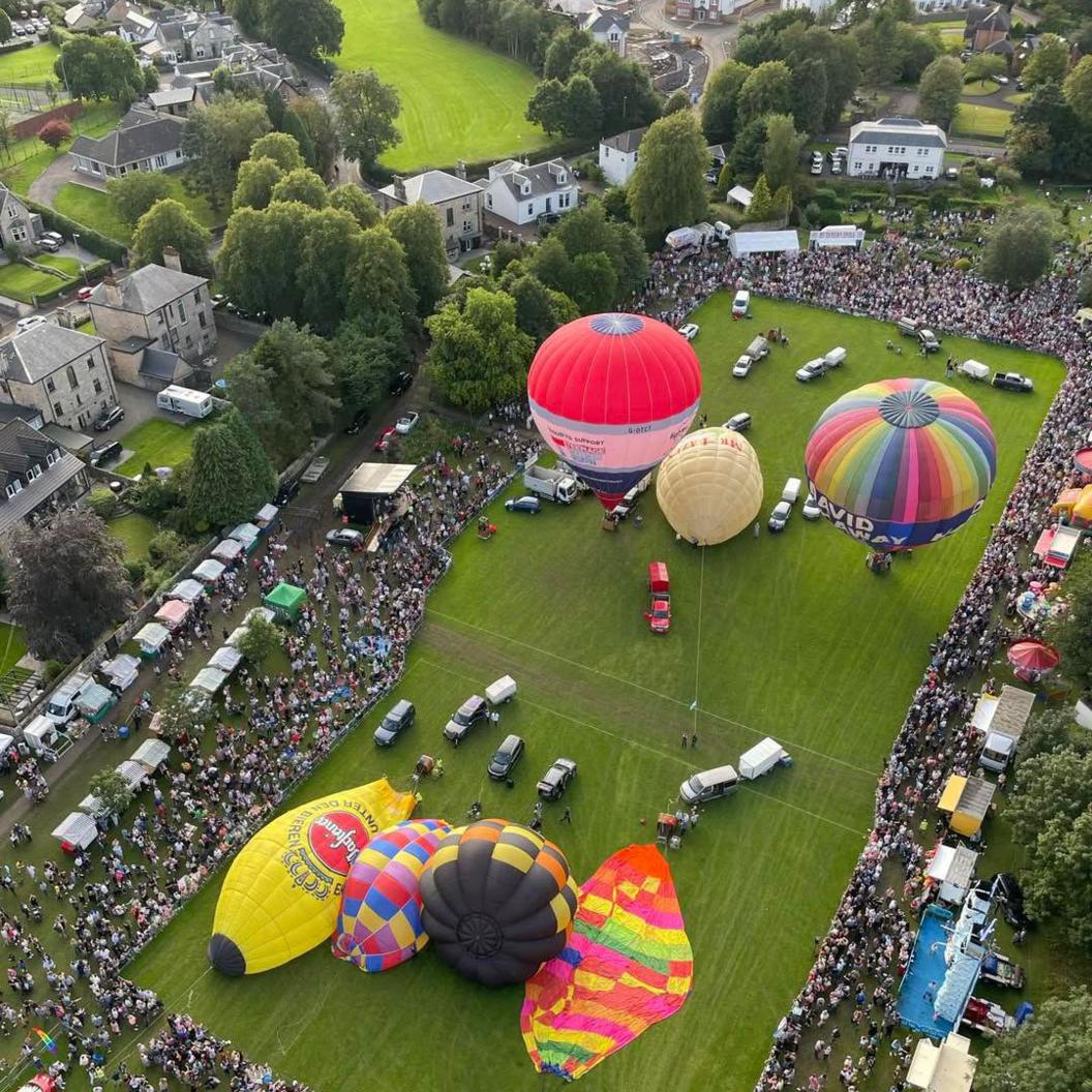 Aerial view as red and yellow hot air balloons are inflated with spectators watching from a distance
