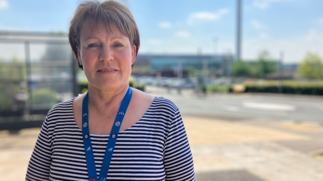 Tracy Bullock standing outside with the hospital blurred in the background. She has short, brown hair and is wearing a white and blue striped top with the straps of a blue landyard around her neck